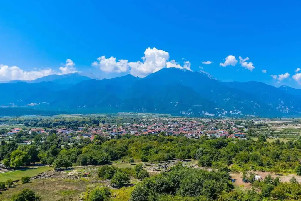 A panoramic landscape of Dion Village in the foreground with the massive, snow-capped peaks of Mount Olympus behind it.