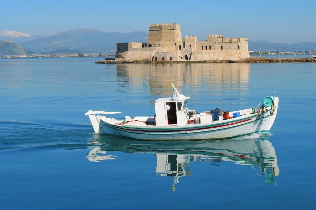 A small, white fishing boat moored in the water directly in front of seaside cafes and tables.