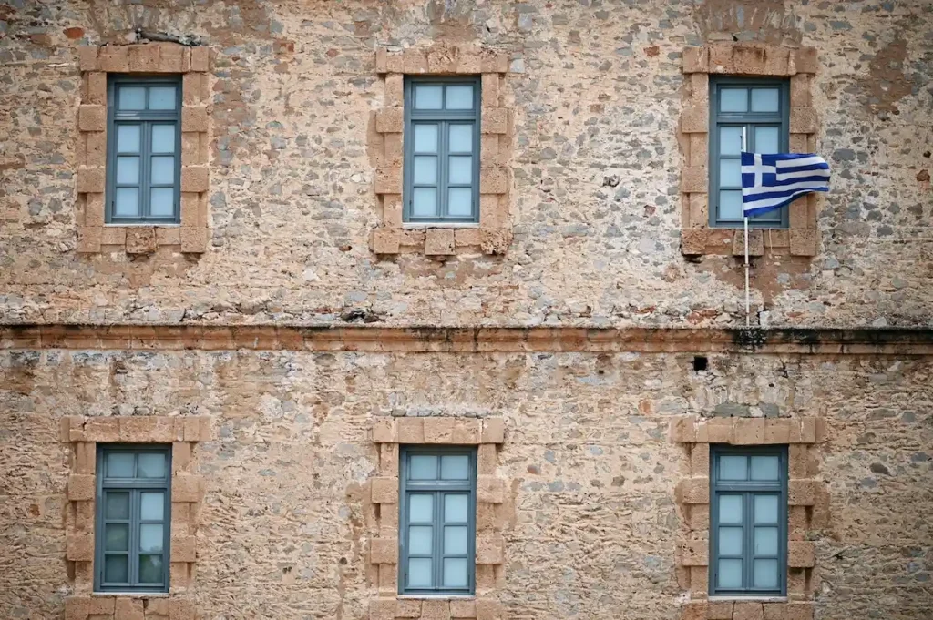 A close-up of a stone wall with a Greek flag waving in the wind.