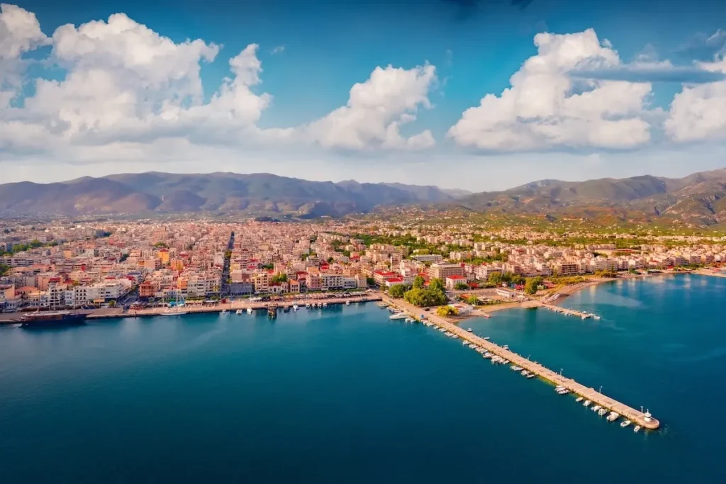 A sweeping aerial view of Kalamata, capturing the operational commercial harbor, the organized marina with yachts, and the long, sandy city beach stretching along the curve of the Messenian Gulf.