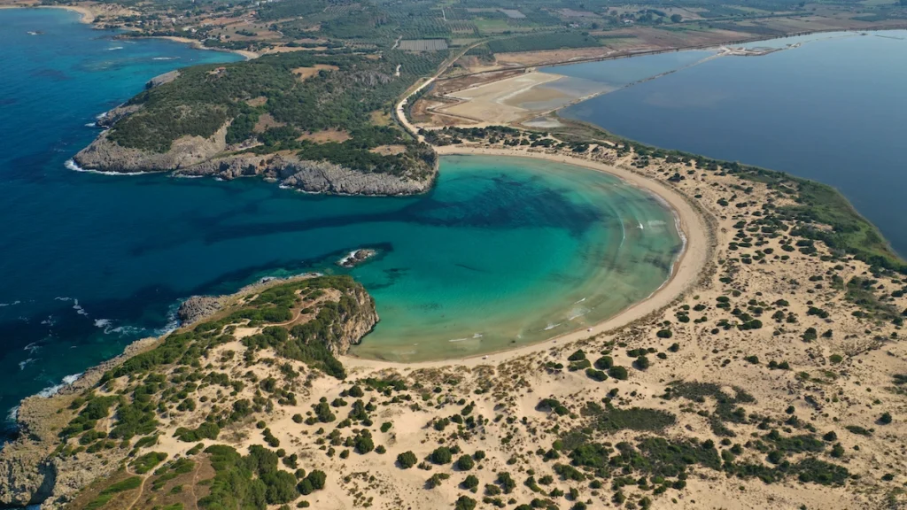 A stunning aerial view of Voidokilia Beach and Lagoon, showing the iconic Omega-shaped bay with turquoise water, a narrow sandy strip, and the dark green lagoon behind it, surrounded by lush vegetation.