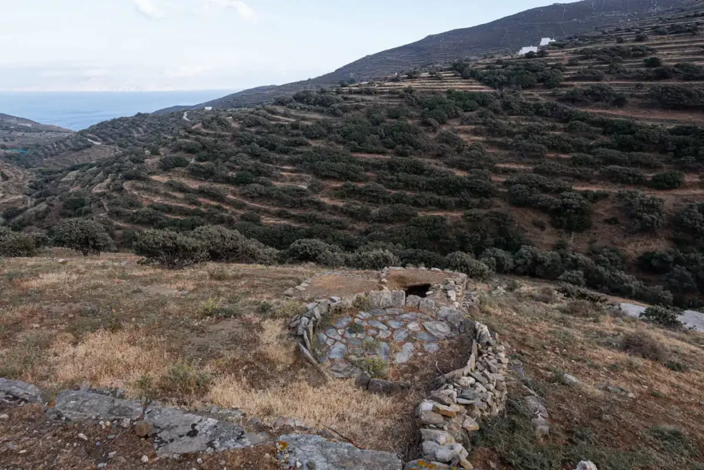 A remote landscape in Tinos featuring an ancient stone threshing floor (aloni) surrounded by wild vegetation with the sea visible in the distance.