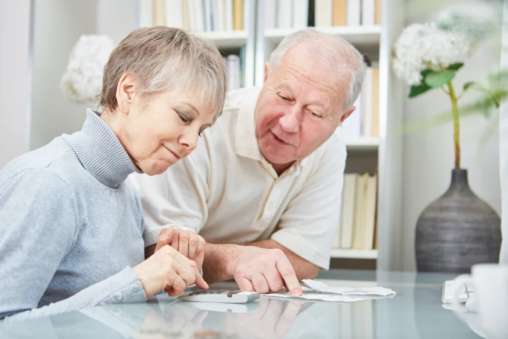 Two retirees sitting at a table, looking at a calculator and paperwork. They are discussing the financial requirements for obtaining a Greek AFM tax number.