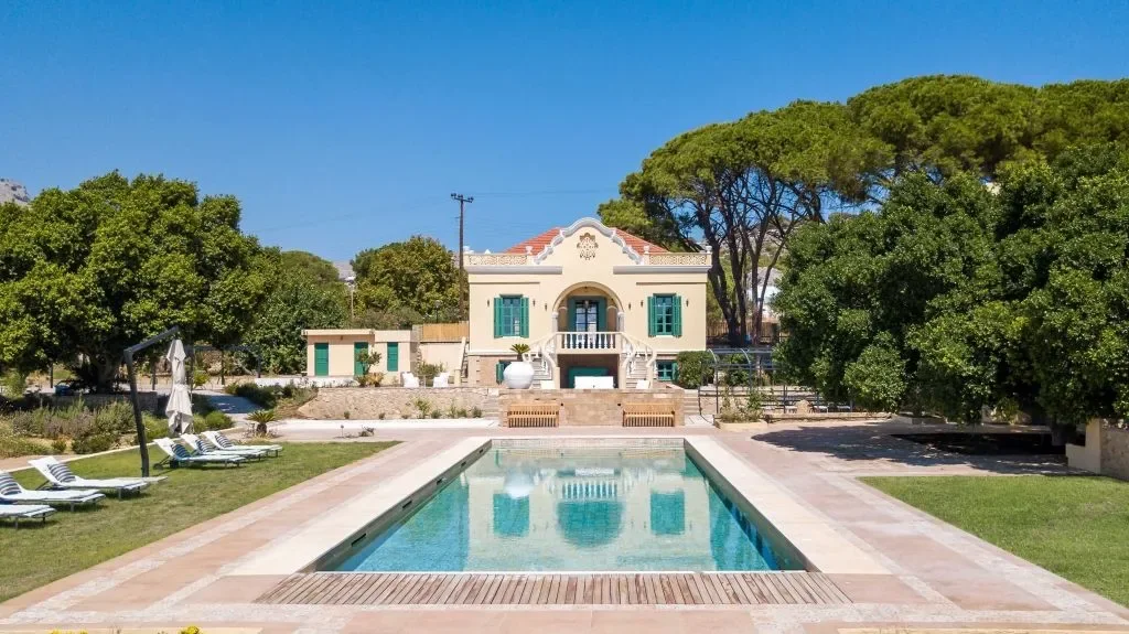 A sprawling, 17th-century historic Lindian mansion featuring high defensive stone walls, a traditional "votsaloto" courtyard with intricate black and white pebble mosaics, and a private swimming pool, set against the backdrop of whitewashed village houses and the towering Acropolis of Lindos.
