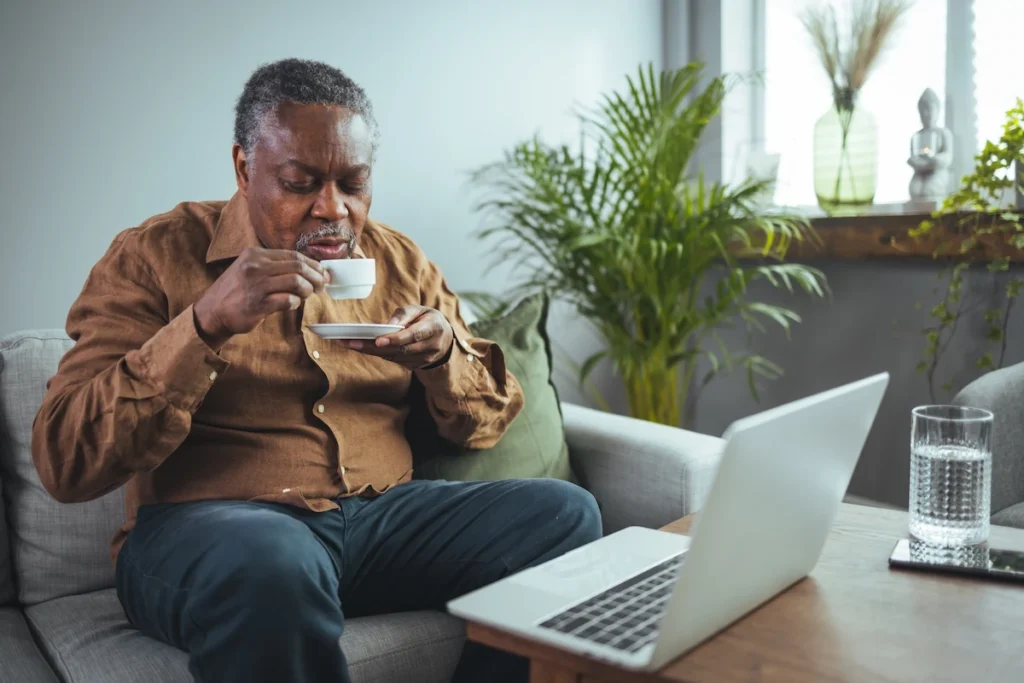 A man relaxing on a sofa with a cup of Greek coffee, managing his residency paperwork on a laptop.