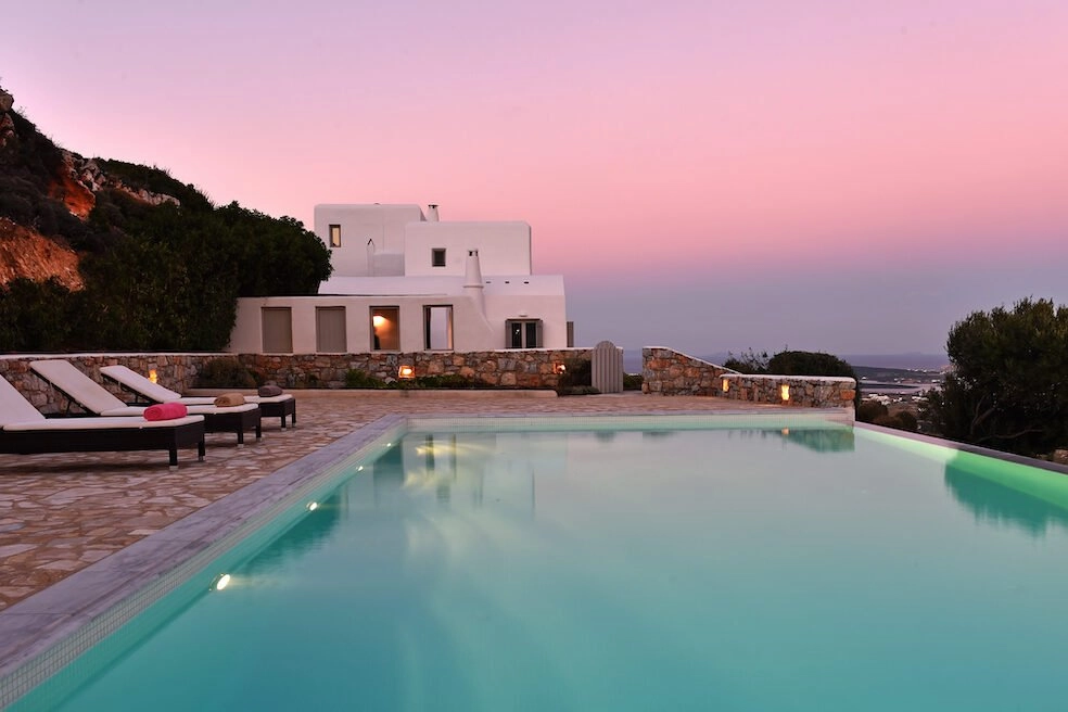 A glowing turquoise swimming pool at a luxury villa in Paros, Greece, set against a vibrant pink and purple sunset sky.