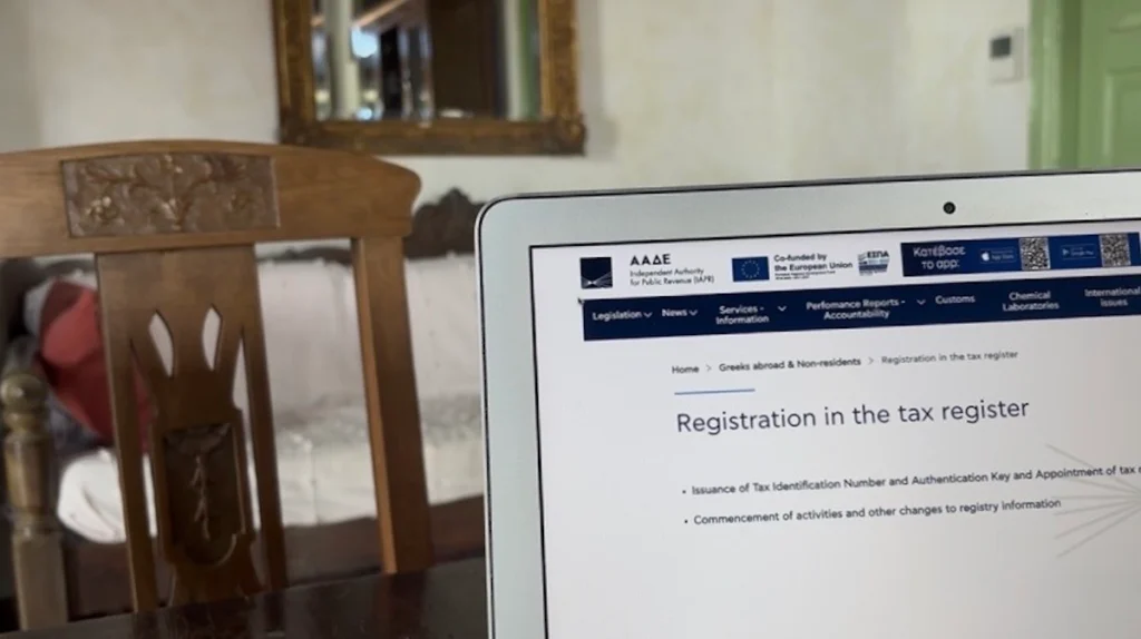 A laptop sits on a wooden table inside a traditional Cycladic house, displaying the Greek Tax Authority (AADE) registration page on its screen against a backdrop of whitewashed walls.