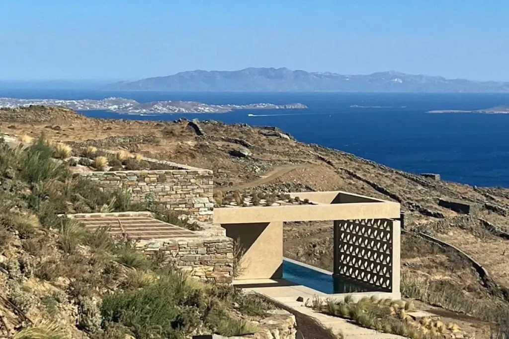 A wide panoramic view from a modern Greek villa terrace looking out over the Aegean Sea toward the silhouettes of Mykonos and Paros.