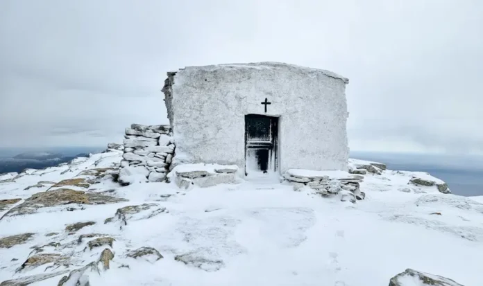 A small, traditional Greek Orthodox chapel sitting in a landscape covered in white snow under a grey sky.