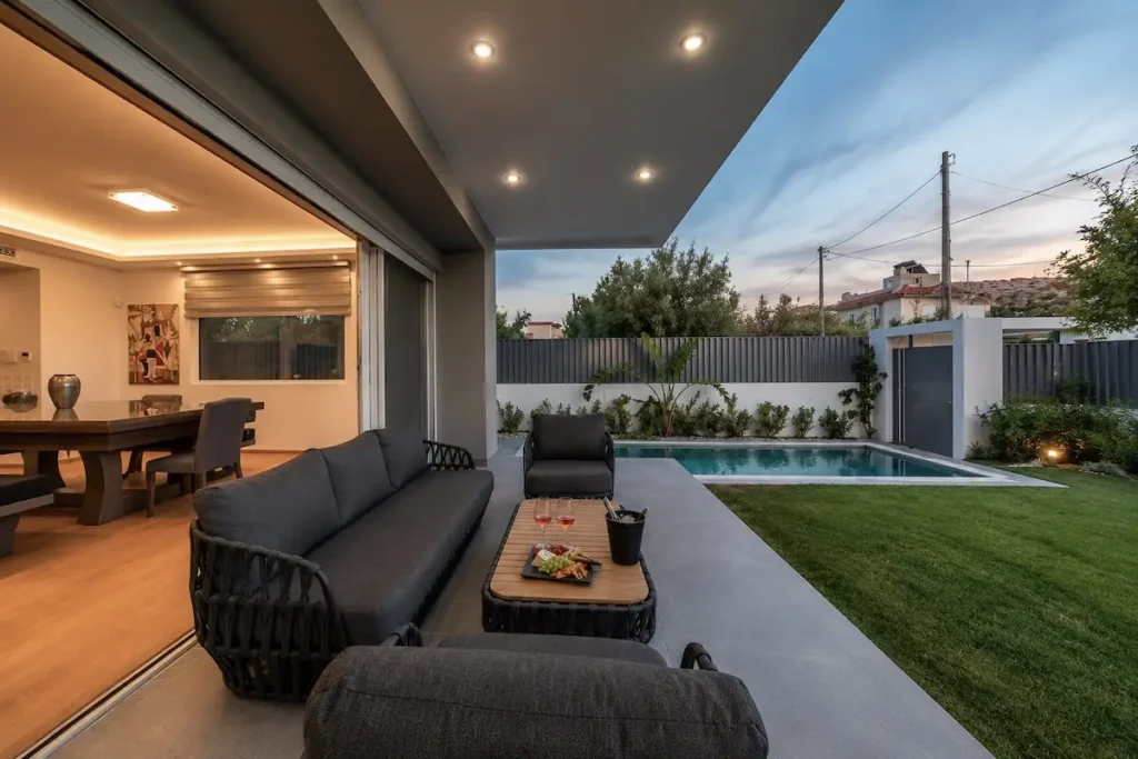 Daytime exterior view of a residential property in a southern Athens suburb, featuring a private swimming pool, landscaped garden, and shaded seating area designed for privacy.