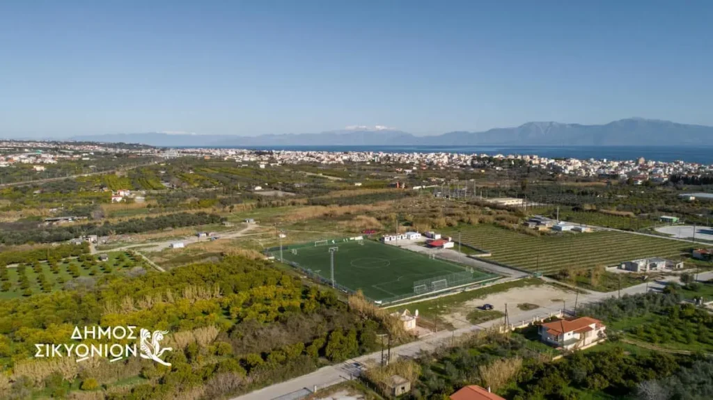 Aerial view near Kiato showing residential land plots, a soccer field, and the surrounding coastal landscape in Corinthia, Greece.