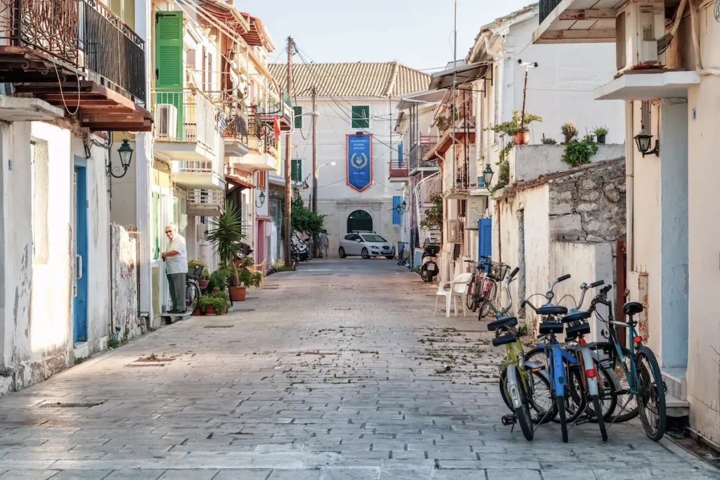 A flat pedestrian street in Lefkada town lined with shops and traditional buildings, featuring residents riding bicycles.