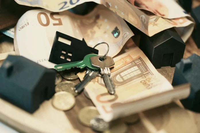 A composition of Euro banknotes, a set of house keys, and a small toy house on a wooden surface.