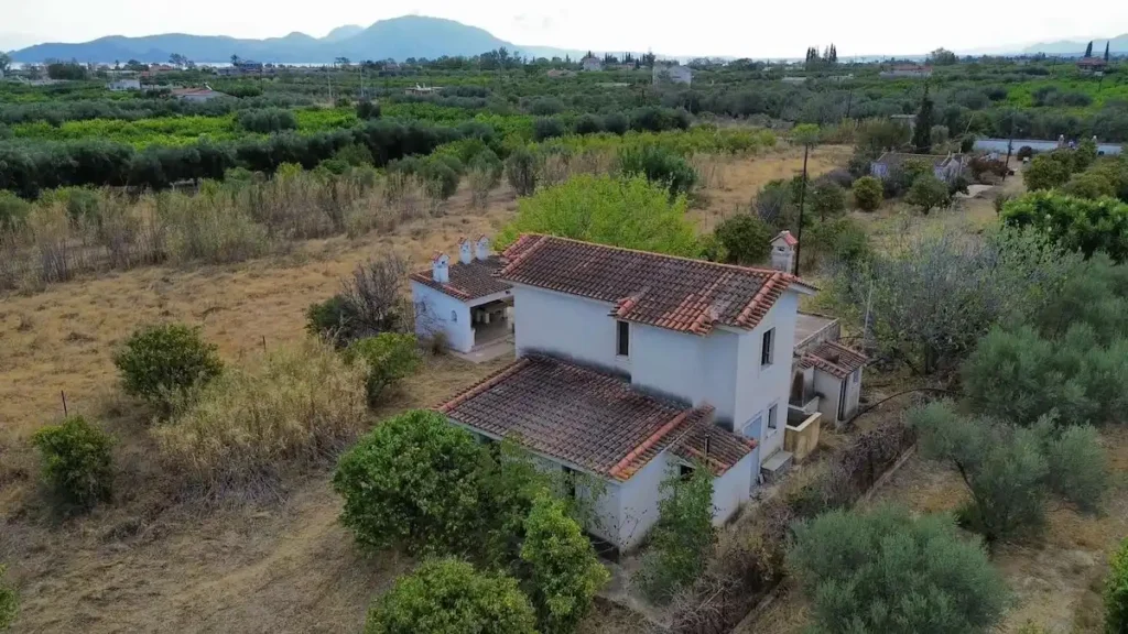 An unfinished 150-square-meter maisonette built in 1990, situated on a large, fenced 6,000-square-meter plot of land in the Agios Ioannis area of Kiato, Greece. An electrical energy pylon is visible in the vicinity.