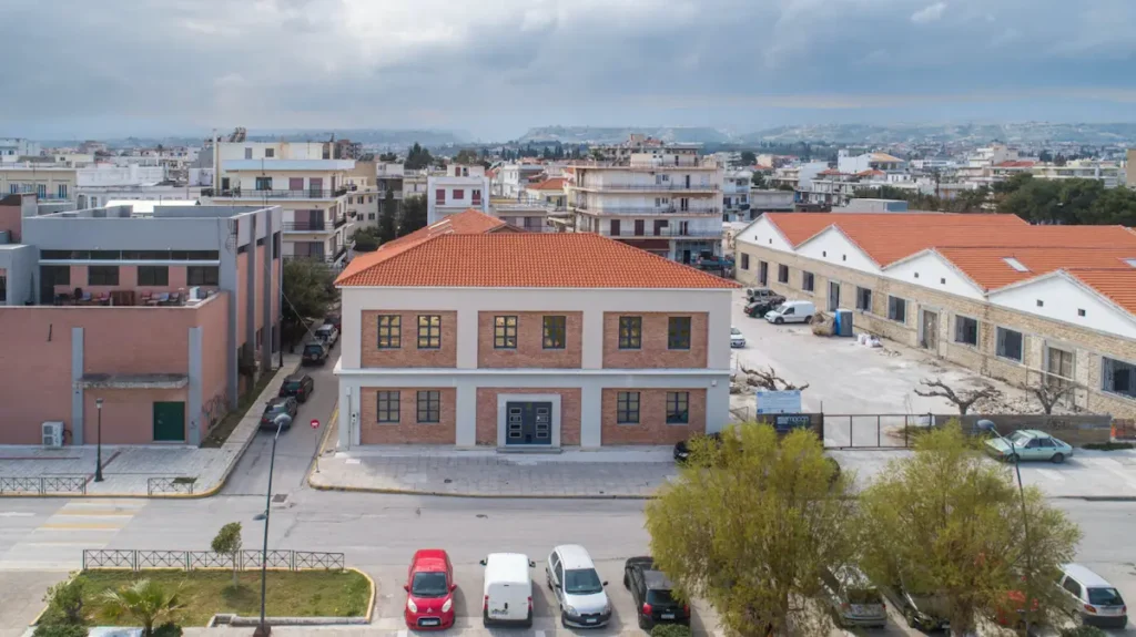 An aerial view of the historic, stone-built municipal cultural complex in Kiato, Greece, showcasing a fully restored auxiliary building that houses the two-floor public lending library alongside five interwar industrial warehouses featuring distinctive, steeply pitched roofs.