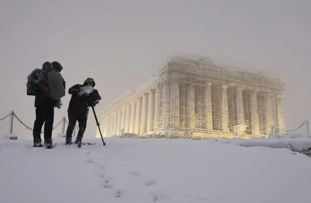 The ancient marble columns of the Parthenon in Athens covered in a light layer of snow with two photographers in the foreground.