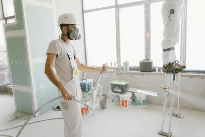 wo construction workers in protective masks and gloves use rollers to paint a section of an unfinished interior wall in a home.