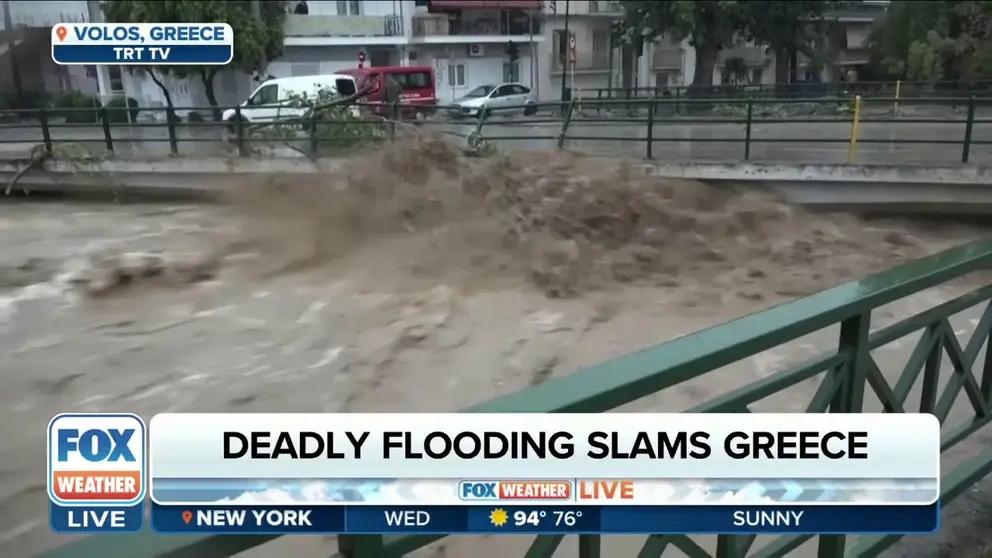 A television news broadcast showing a flooded city street with cars partially submerged in brown water and debris.