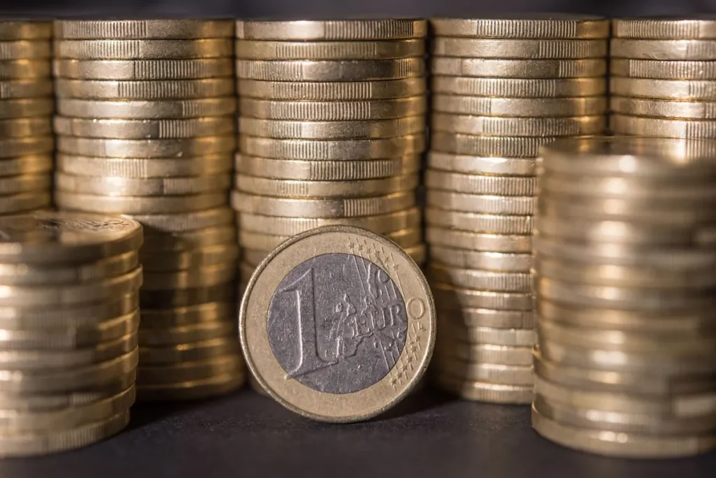 A single one-euro coin stands perfectly balanced on its edge in the foreground, with a blurred wall of stacked euro coins creating a solid background.