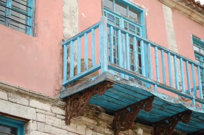 The upper floor of a historic neoclassical building featuring an exposed stone lower level, a faded pink upper level, and weathered blue-turquoise wooden window frames and balcony doors. The ornate, artistic iron supports of the balcony show visible rust and lack of maintenance.