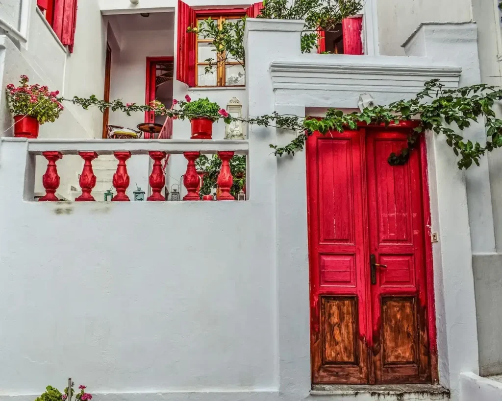 A traditional two-story white house in Chora, Skopelos, featuring a vibrant red wooden door and window frames, potted red geraniums, and a lush green climbing plant framing a private entrance that leads to an interior staircase and balcony.