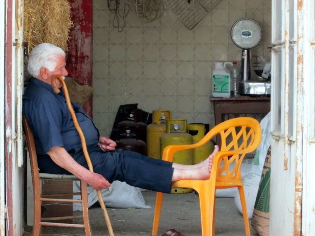 An elderly Greek man resting on a wooden chair at the entrance of a traditional village grocery store.