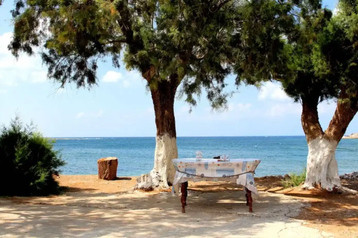A rustic wooden dining table and chairs shaded by trees on a sunny, quiet sandy beach in Phalasarna, Greece.