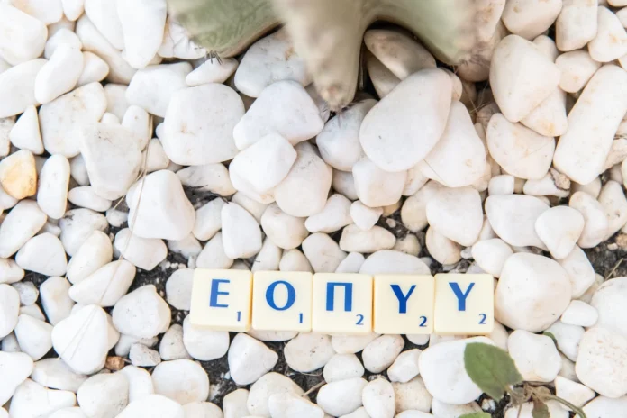 Wooden Scrabble tiles spelling out the acronym EOPYY, arranged neatly on a bed of smooth, white decorative pebbles.
