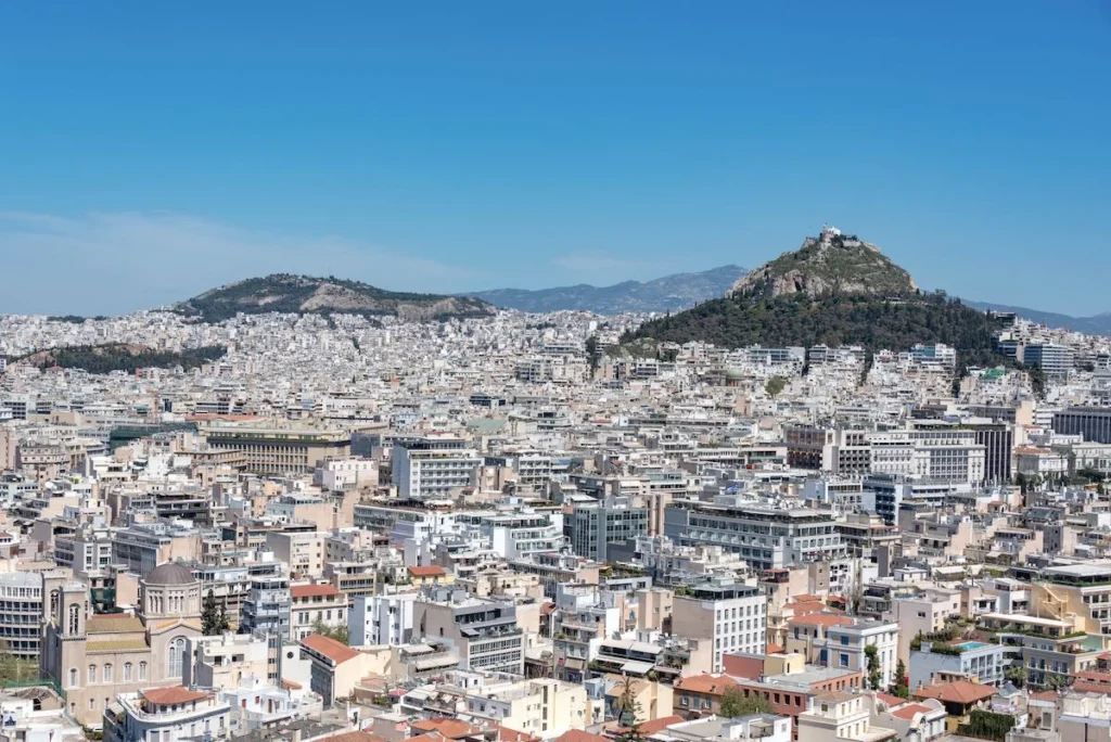 A sprawling aerial view of the dense Athens cityscape showing crowded concrete apartment buildings under a clear blue sky, with the peak of Lycabettus Hill rising in the background.