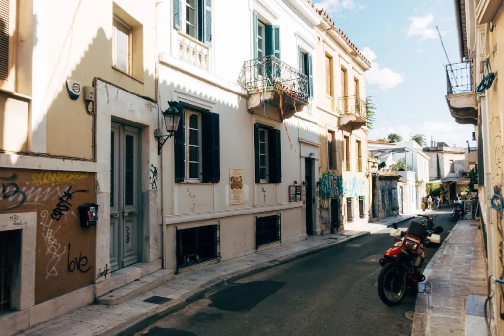 A row of neoclassical Athens houses, including one needing renovation and one freshly painted, with graffiti tags on the walls and scooters parked by the curb.