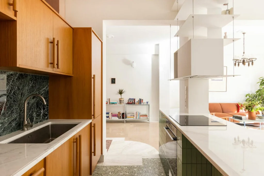 A high-end renovated mid-century modern Athens apartment interior featuring sleek wooden built-in closets that conceal major appliances, vibrant green wall tiles, and minimalist design elements.
