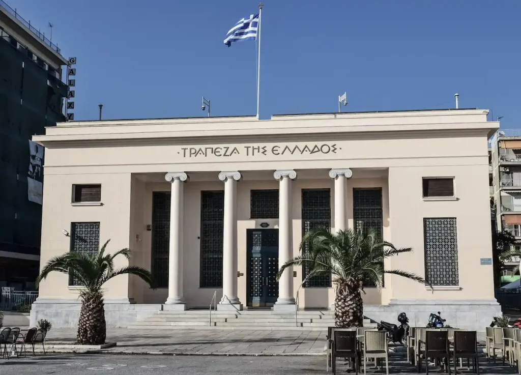 Neoclassical exterior of the Bank of Greece branch in Volos with beige walls and tall stone columns.