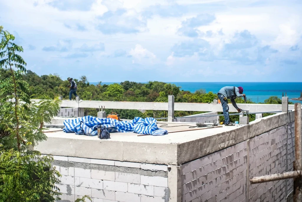 Two construction workers on the roof of a house under construction, with the sea in the background.