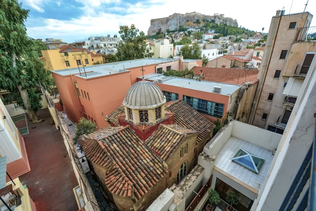 Athens view looking up toward the Acropolis stone walls, featuring a traditional church with red clay tile roofing surrounded by peach, ochre, and modern apartment buildings.