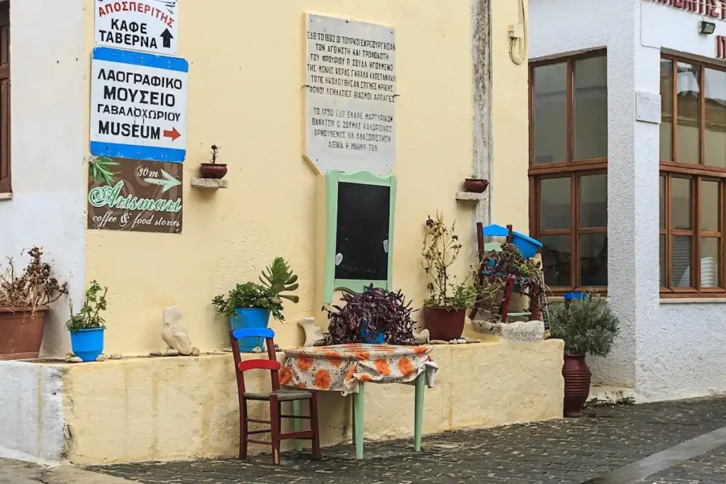 A rustic outdoor table with a floral tablecloth and a traditional kafeneio chair outside the Gavalochori Folklore Museum.