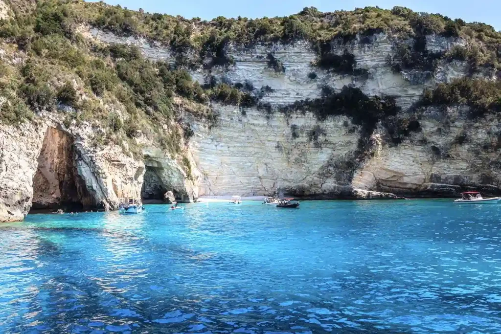 Tall limestone cliffs dropping into deep blue sea with small boats moored near the shore on Paxoi.