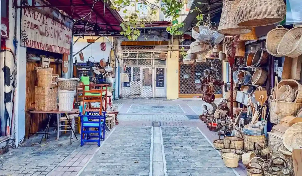 A colorful pedestrian street in Thessaloniki lined with traditional shops displaying woven baskets and outdoor chairs.