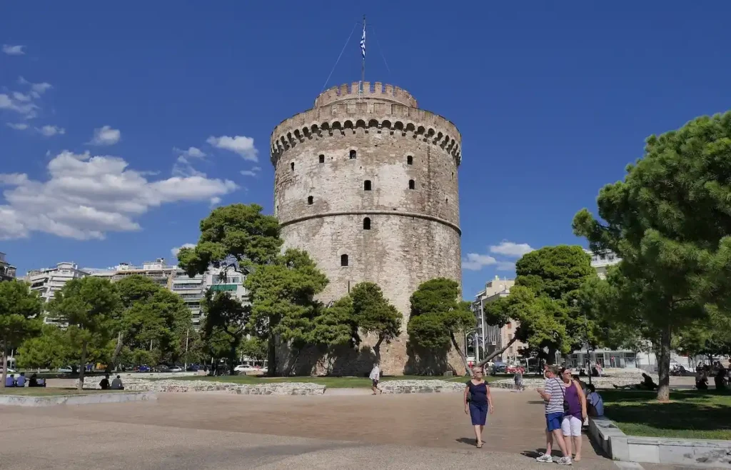 The historic White Tower of Thessaloniki on a sunny day with pedestrians dressed in summer clothes walking nearby.