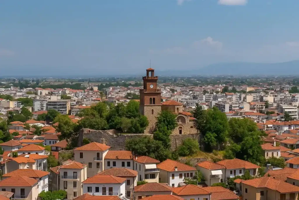 A panoramic view of Trikala city featuring the iconic Clock Tower, historic old town buildings, and a modern cityscape stretching toward a distant valley.