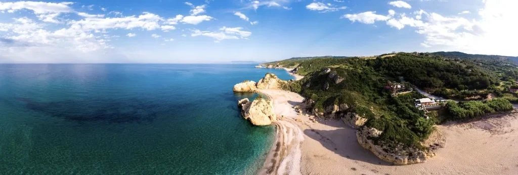 A tranquil beach just outside Volos, featuring clear blue waters, soft golden sand, and rugged rocky cliffs under a bright Mediterranean sky.