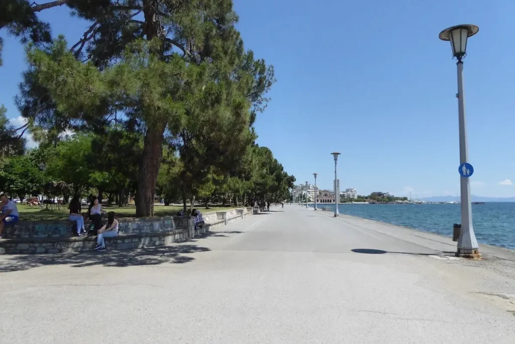 A peaceful view of the wide, paved pedestrian coastal promenade in Volos, lined with shady trees, park benches, and a classic metal lamppost overlooking the water.