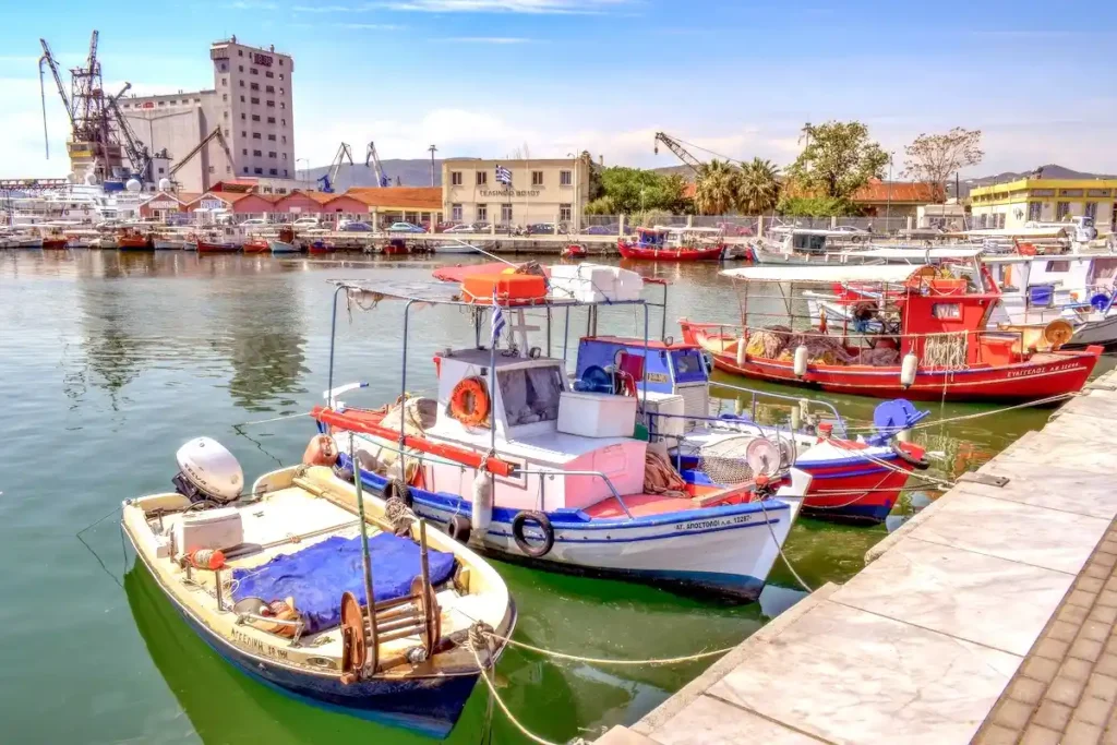 Vibrant, colorful traditional Greek fishing boats moored at the Volos harbor, with the historic stone Custom House (Teloneio) building visible in the background under a clear sky.