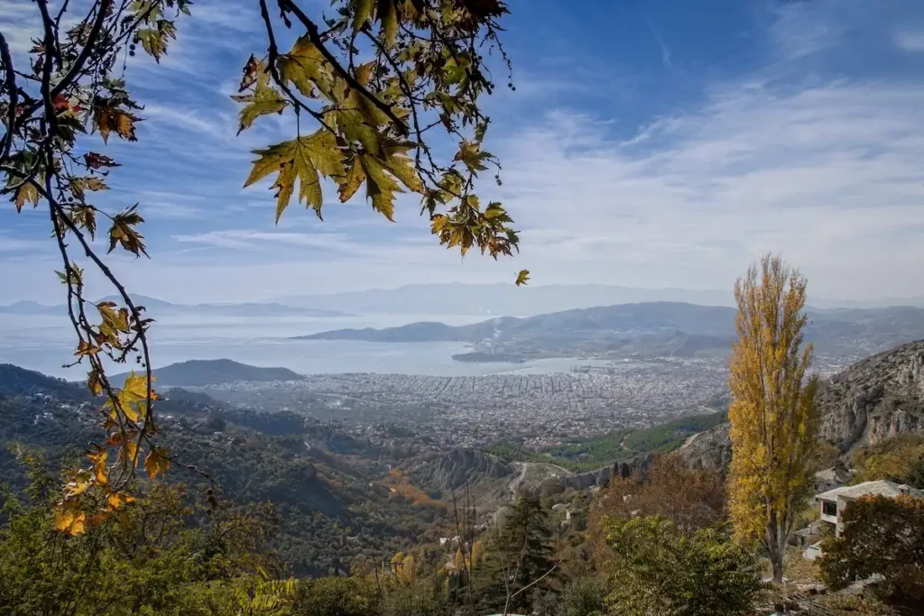 Panoramic, wide-angle view from high up on Mount Pelion looking down at the sweeping blue bay of the Pagasetic Gulf, the sprawling city of Volos, and the surrounding green nature.