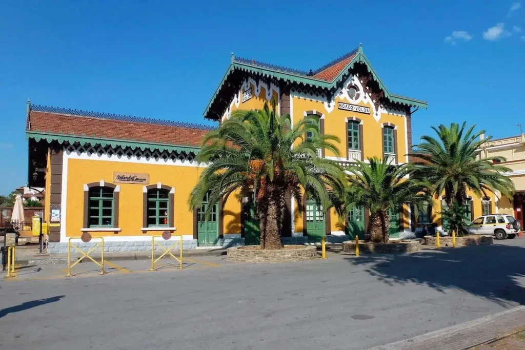 The historic Volos Train Station building, featuring traditional yellow-ochre walls, elegant green doors, and green window frames, framed by green palm trees. Designed by Evaristo de Chirico.