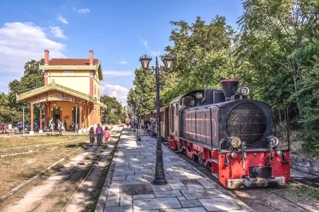The historic black steam train of Pelion with bright red metallic detailing, sitting on narrow tracks surrounded by lush green trees on the mountain.