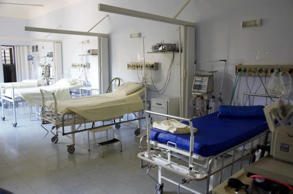 An empty, clean hospital ward containing four medical beds with white linens, separated by privacy curtains.