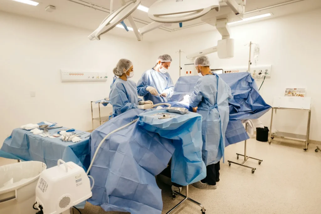 Three surgeons in blue sterile scrubs, masks, and caps stand over a surgical table under bright medical lights in an operating room.