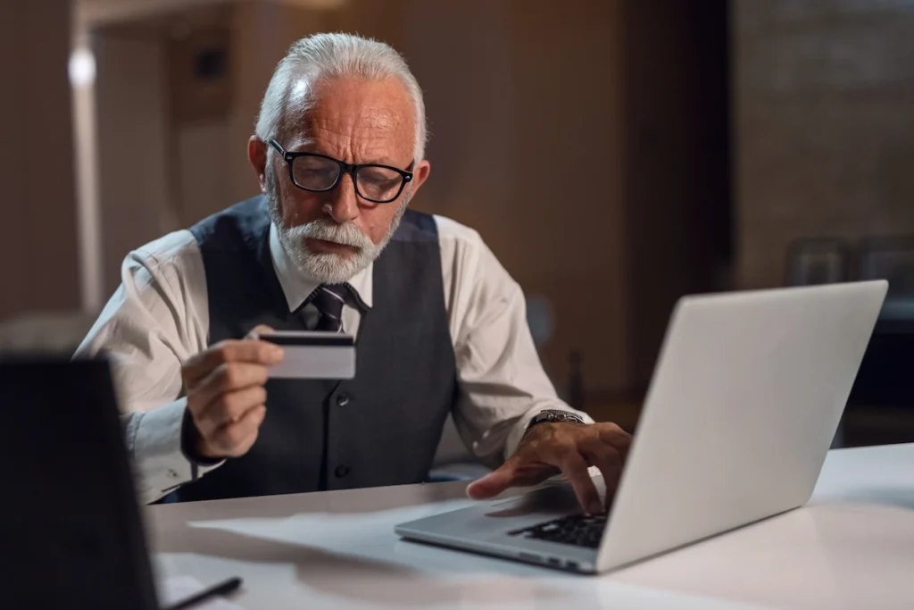 A senior man with glasses and a grey vest holds a credit card while using a silver laptop.
