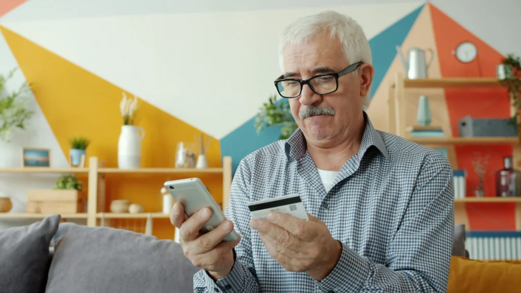 A smiling mature man using a smartphone and bank card for online banking inside his home.