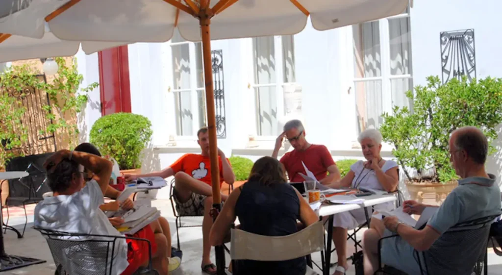 A group of adult language students seated at outdoor tables conversing in the stone courtyard of The Athens Center surrounded by greenery and Mediterranean architecture.